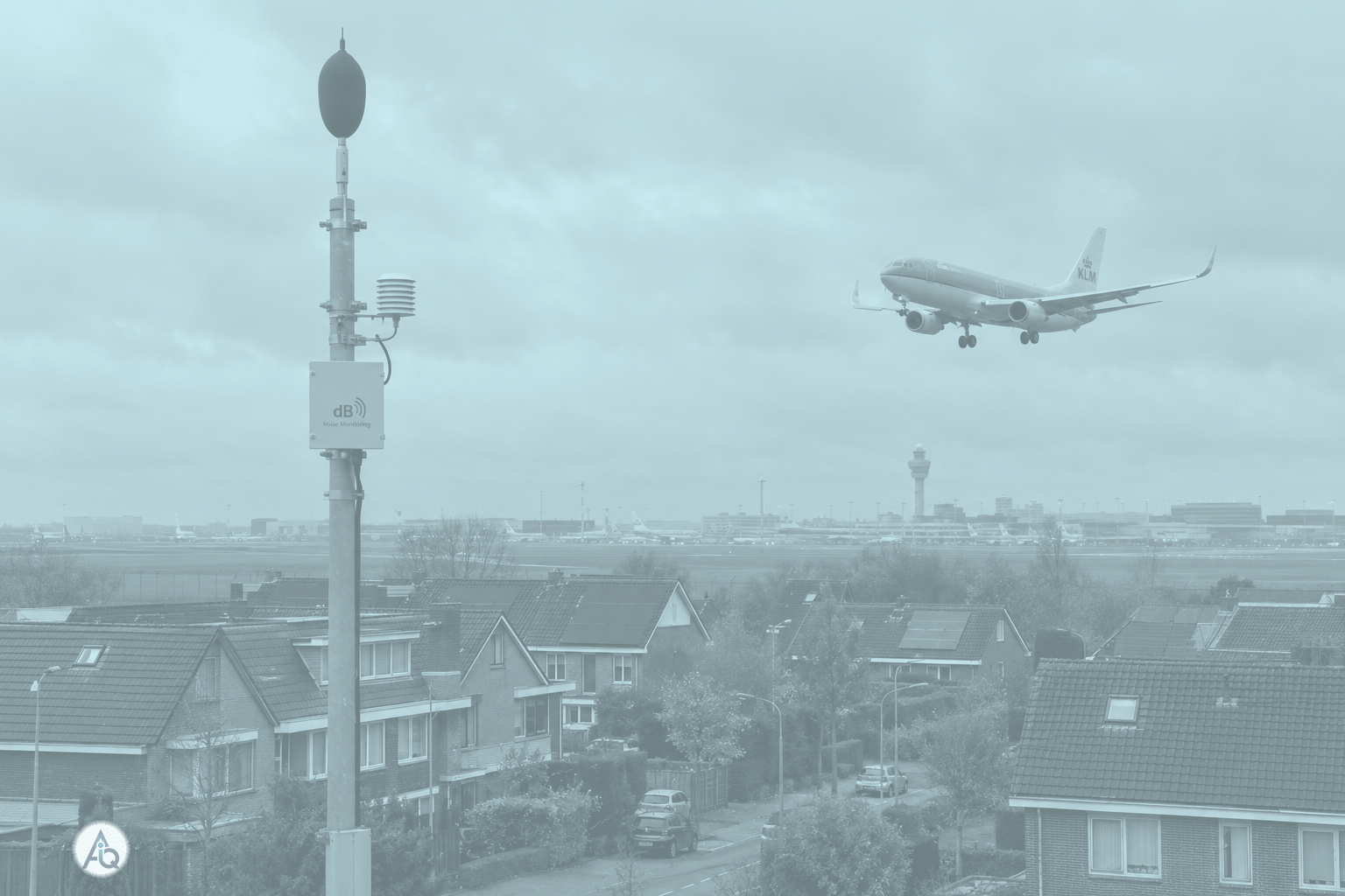 Woonwijk aan de rand van een vliegveld met een geluidsmeter-mast op de voorgrond en een naderend vliegtuig